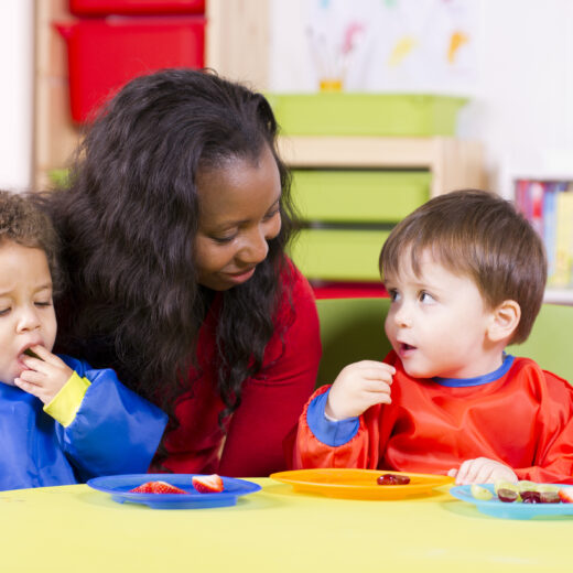 Teacher talking with two toddlers eating fruit.