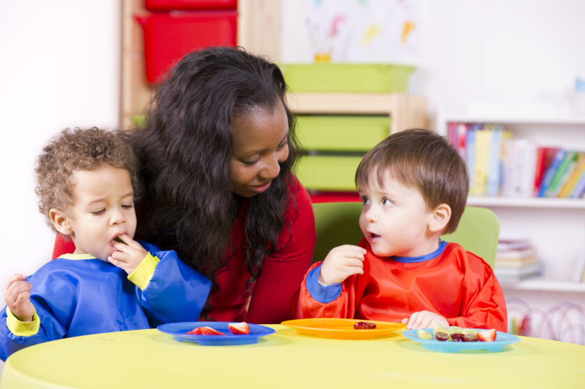 Teacher talking with two toddlers eating fruit.